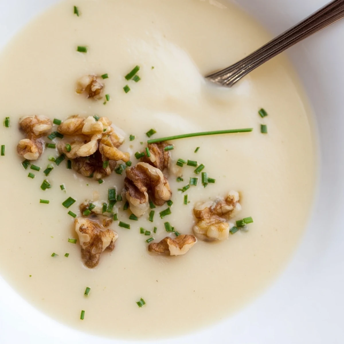 A bowl of creamy parsnip and pear soup topped with toasted walnuts, chives, and a drizzle of cream.