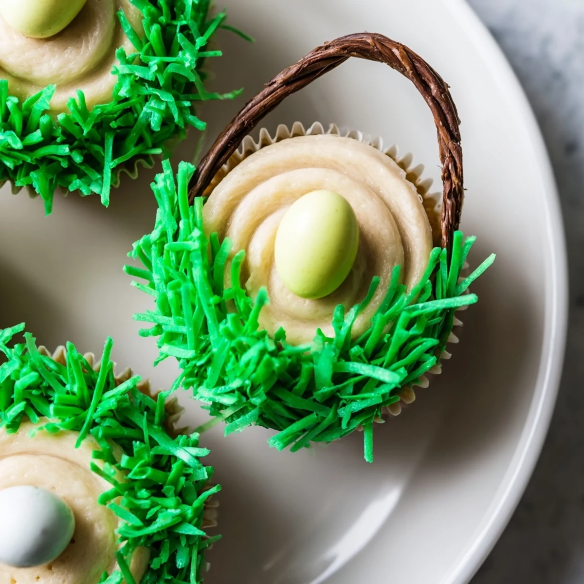 Two Easter Basket Cupcakes with Coconut Grass feature licorice basket handles and pastel candy eggs nestled in vibrant green frosting.