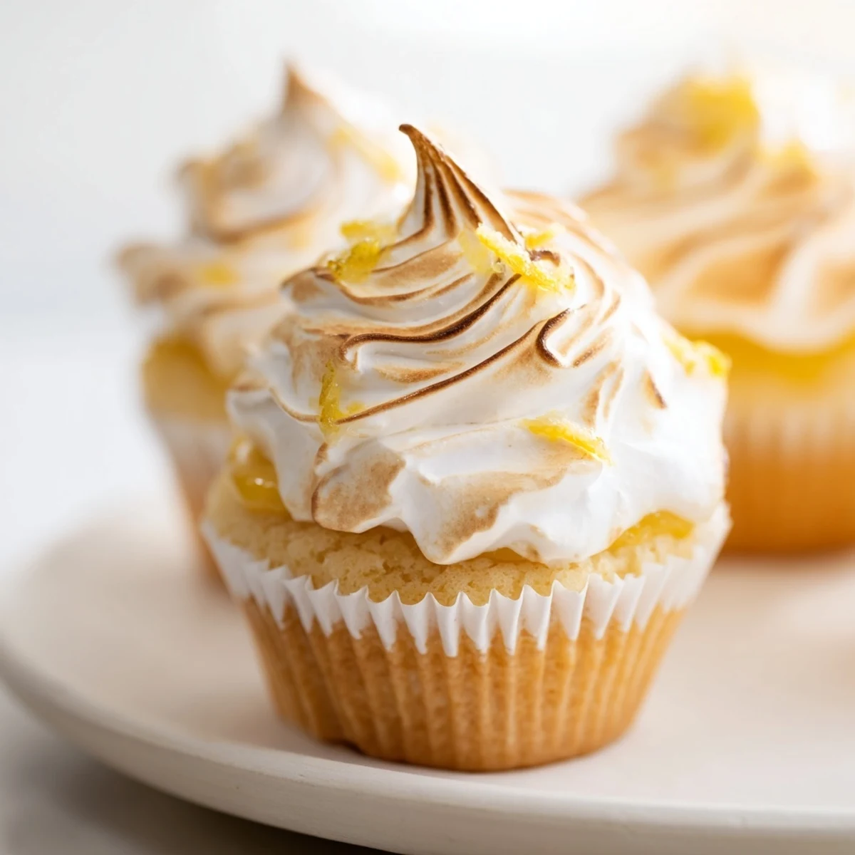 A close-up view of Lemon Meringue Cupcakes showing billowy meringue peaks and yellow curd filling beside a glass of milk.  
