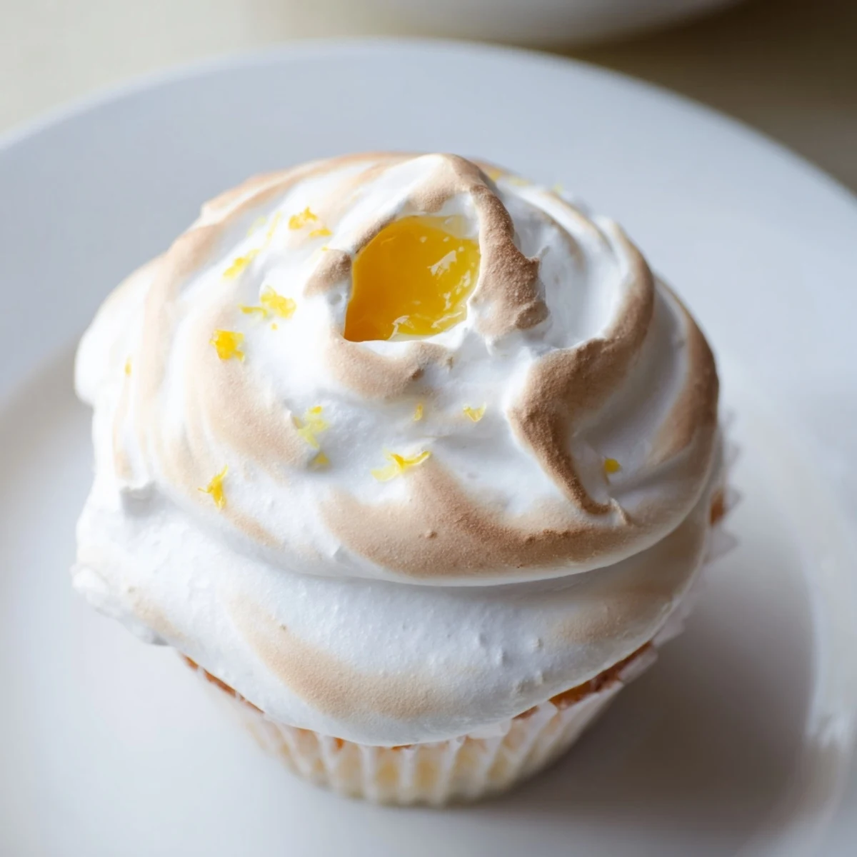 Twelve lemon meringue cupcakes sit on a white plate, featuring golden toasted meringue swirls and a vibrant yellow curd visible in the cross-section.