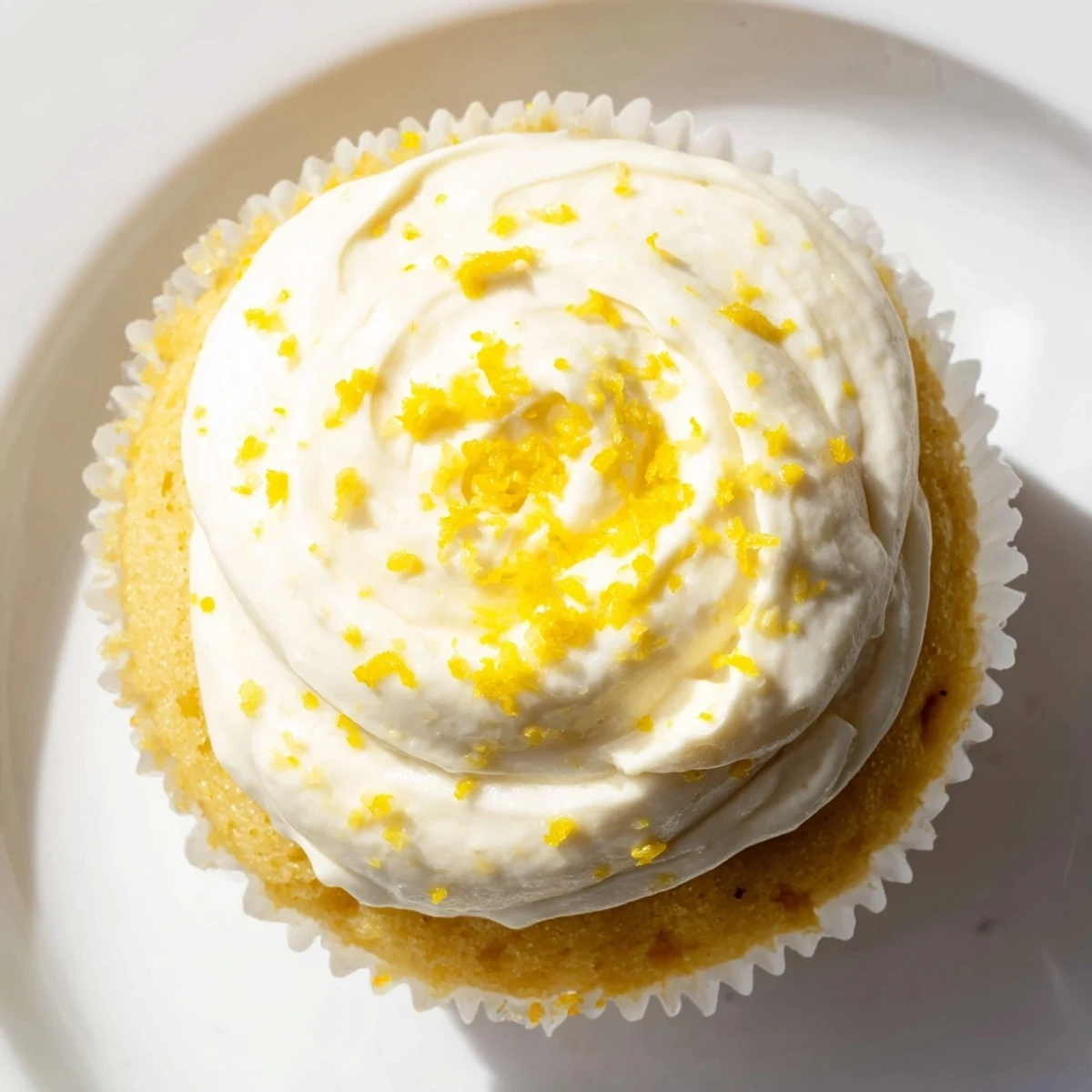 Twelve golden Limoncello cupcakes sit on a white plate with creamy frosting swirls and fresh lemon zest garnish.  