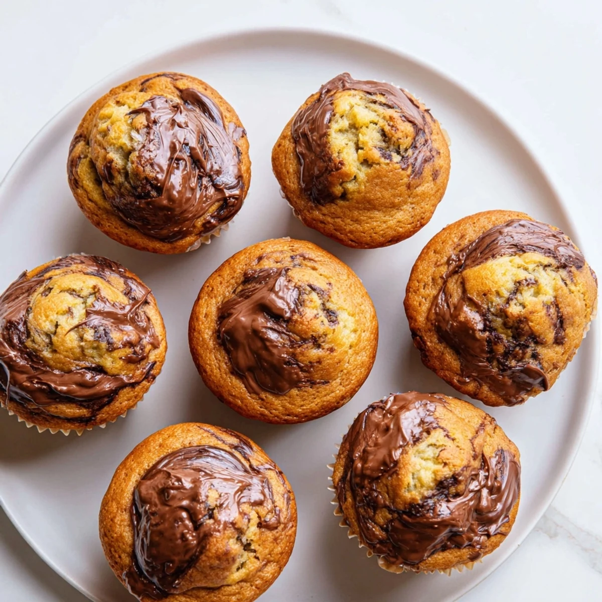 Golden brown Nutella banana muffins topped with a hazelnut chocolate swirl on a white plate