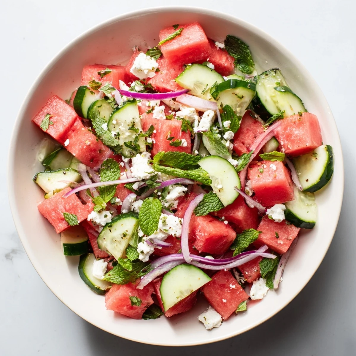 Fresh watermelon feta salad bowl with juicy red melon cubes, crumbled white feta cheese, cucumber, and mint leaves drizzled with olive oil dressing