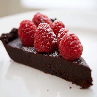 A fresh Decadent Dark Chocolate Tart with Raspberries is served on a white plate, dusted with powdered sugar and ripe red berries.